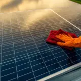 Ręka w pomarańczowej rękawiczce czyści panele fotowoltaiczne czerwoną ściereczką. Czyste panele to więcej słońca i energii.