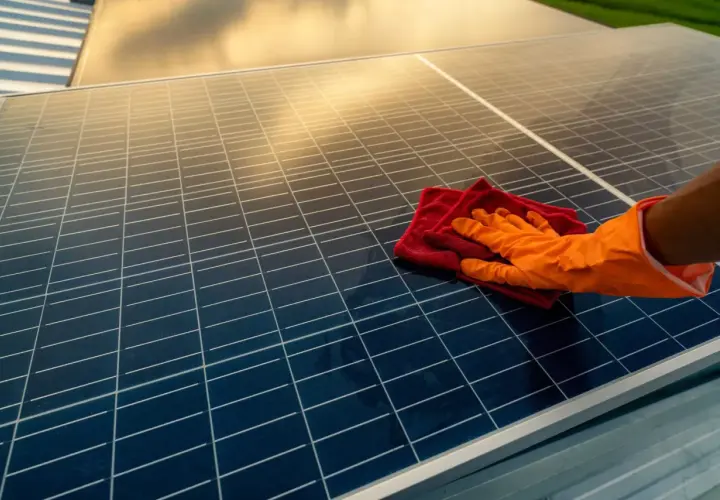Ręka w pomarańczowej rękawiczce czyści panele fotowoltaiczne czerwoną ściereczką. Czyste panele to więcej słońca i energii.