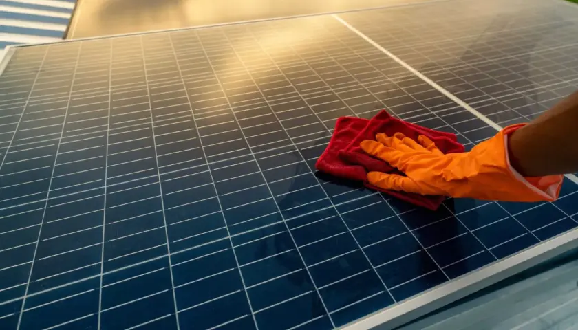 Ręka w pomarańczowej rękawiczce czyści panele fotowoltaiczne czerwoną ściereczką. Czyste panele to więcej słońca i energii.